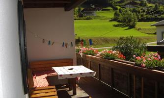 A balcony with a wooden bench and a table. In the background, green meadows and residential houses can be seen, surrounded by mountains.