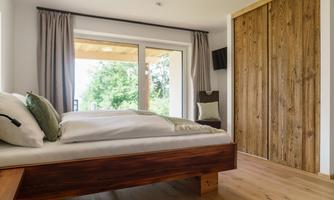 A modern bedroom with a wooden bed and large window doors. The room is bright and friendly, with a view of the greenery.
