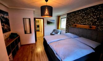 A modern bedroom with a large double bed and a cozy atmosphere. The walls are partly decorated with floral patterns, and there is a mirror as well as a chair in the room.