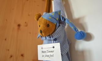 A plush bear in a blue pajamas is hanging on a wall. It is wearing a sign that says "My room is now free!".