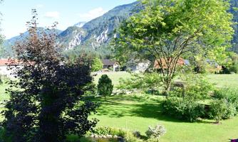 A picturesque landscape with lush green, trees, and a view of the mountains. In the background, some houses are visible.