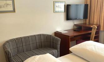 A cozy hotel room with a sofa, a desk, and a television. The room has light curtains and simple, modern furniture.