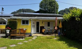 A small, yellow house with a well-maintained garden. There is an outdoor seating area and umbrellas to provide shade.