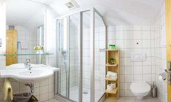 A modern bathroom with a glass shower and light tiles. There is a sink and a wooden shelf with towels and toiletries.