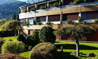 A modern building with wooden cladding and large windows surrounded by green gardens. The sky is blue and the landscape appears calm and inviting.