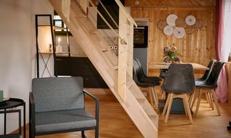 A cozy living space with a wooden wall and an open kitchen. A dining area with a table and chairs, along with modern furniture, creates an inviting atmosphere.