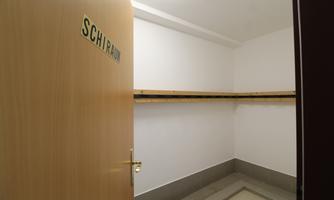 A room with an open door, labeled "SCHIRAU." The walls are white and there is a wooden shelf on the wall.