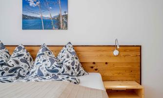 A cozy bedroom with a wooden bed and elegant cushions. A picture of a lake under a blue sky hangs on the wall.