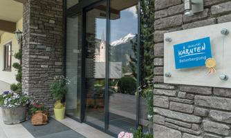An inviting entrance to an accommodation with modern windows and a stone wall. In the background, green plants and snow-covered mountains are visible.