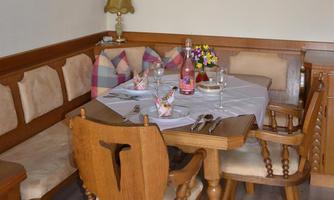 A cozy dining table with wooden chairs and a decorative tablecloth. On the table are glasses, cutlery, and a vase of flowers.