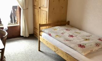 A simple bedroom with a single bed, a wooden wardrobe, and a table. The window lets in natural light and creates a bright atmosphere.