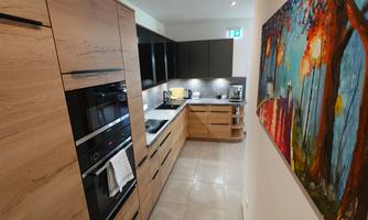 A modern kitchen with wooden cabinets and sleek appliances. A colorful painting of a landscape hangs on the wall.