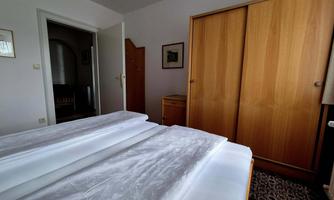 A simple bedroom with two beds and a wooden wardrobe. The walls are light-colored and there is a carpeted floor.