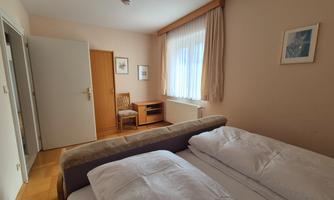 A cozy bedroom with two beds and a table. Large windows provide plenty of daylight.