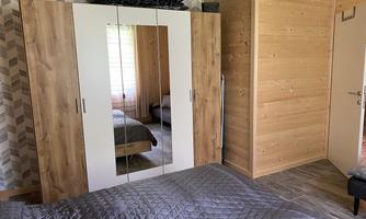 A cozy bedroom with a large wooden wardrobe and a bed with a gray bedspread. The walls are in warm wood tones, creating an inviting atmosphere.