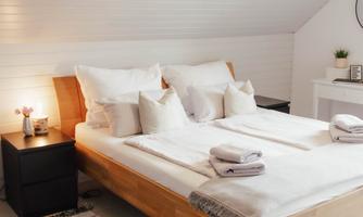 A cozy bedroom with a large wooden bed and many pillows. The room has light walls and a modern carpet.