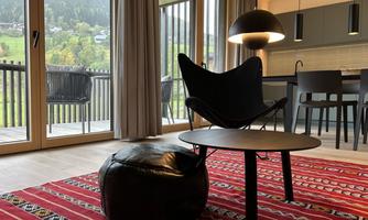 A modern living room with a black armchair, a round table, and a stool. Large windows offer a view of the green landscape outside.