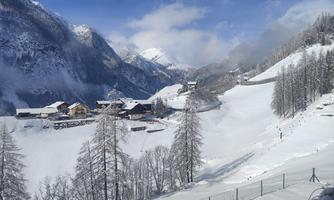 A snow-covered mountain landscape with several cottages in the valley. The surroundings are calm and picturesque, surrounded by tall mountains and a clear sky.