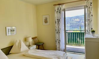 A cozy bedroom with a view of the landscape. The room is bright and friendly with pretty curtains and a small seating area.