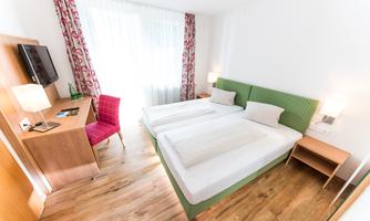 A cozy hotel room with two beds and wooden flooring. The room is bright and modernly furnished, with a desk and a television.