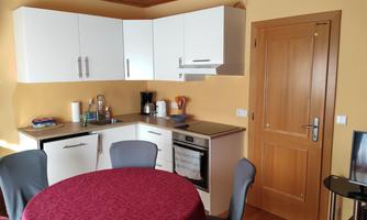 A modern kitchen with white cabinets and a round, red-covered table. The walls are painted in a warm yellow tone and the door is made of wood.