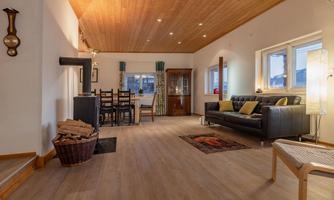 A cozy living room with a wooden ceiling and large windows. There is a black sofa, a dining table, and a fireplace.
