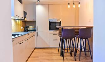 A modern kitchen with a light wood floor and elegant furniture. The bar is equipped with tall stools, and the lighting creates a cozy atmosphere.