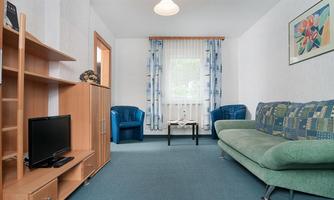 A cozy living room with a green sofa and blue chairs. A television sits on a wooden shelf, and curtains let in soft light.