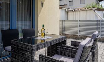 A cozy outdoor area with a table and chairs made of rattan. On the table sits a bottle of wine and a glass.