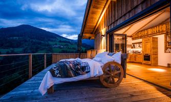 A stylish terrace with a wooden bed and a view of the mountain landscape. The warm lighting creates a cozy atmosphere.