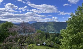 A picturesque landscape with green forests and a blue lake. In the background, there are mountains and a clear sky with white clouds.
