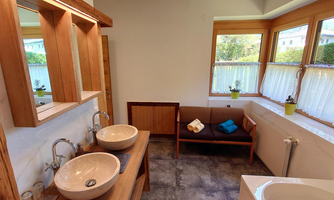 A modern bathroom with natural wooden elements and two round washbasins. Large windows provide plenty of daylight and a view of the greenery.