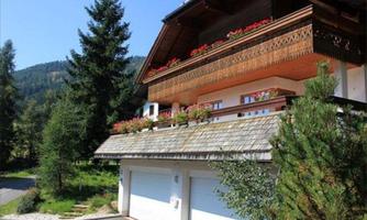 A beautiful house with a balcony and flowers. In the background, there are trees and mountains.