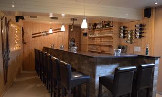 A cozy bar with wooden interiors and a long counter. In the background, there are shelves with drinks and warm lighting.