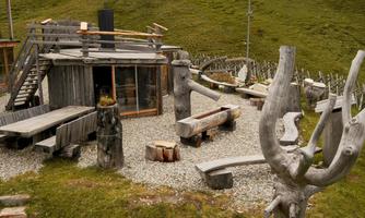 A cozy wooden cabin with external wooden seating. The surroundings are characterized by green grass and rocks.