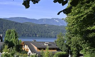 A peaceful lake surrounded by mountains and trees. In the foreground, some houses and green plants are visible.