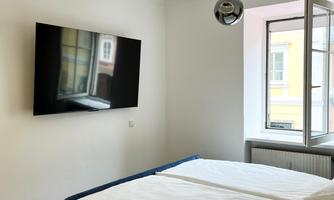 A modern bedroom with a double bed, a television on the wall, and a window. The room is bright and simply designed.