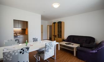 A modern living room with a dining area and a small kitchen. The furniture is in neutral colors, and the room feels bright and inviting.