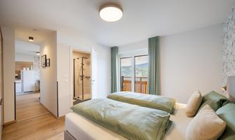 A modern bedroom with two separate beds and striking green bedspreads. Large windows provide a view of the terrace and the area with a bathroom behind it.