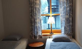 A cozy bedroom with two single beds and a small table. The window lets in plenty of daylight and is adorned with curtains.