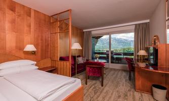 A modern hotel room with wooden elements and a view of the mountains. The room is bright and comfortably furnished, with a large window and a cozy seating area.