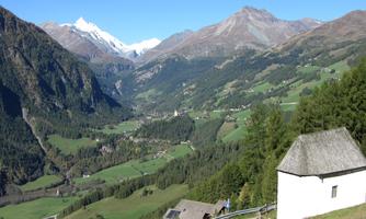 A picturesque mountain landscape with snow-capped peaks and a green valley. In the foreground stands a small, white building.