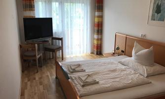 A simple room with a double bed, a television, and two chairs. Large windows let in plenty of daylight.