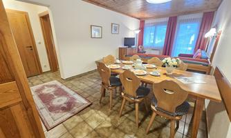 A cozy dining area with a large table and chairs. In the background, there is a living room with a sofa and a television.