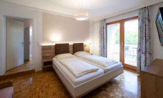 A cozy bedroom with two beds and hardwood flooring. There are large windows that lead to a balcony and let in plenty of daylight.
