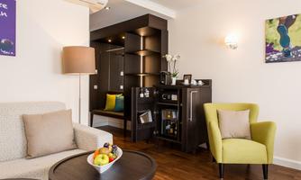A cozy living space with a sofa, an armchair, and a small table. In the background, there is a minibar and a spot to relax.