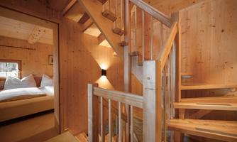 A bright wooden staircase in a cozy interior. In the background, there is a bedroom with a large bed and soft pillows.
