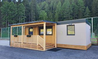 A modern wooden structure with a veranda and large windows. Surrounded by green trees and a clear mountain backdrop.
