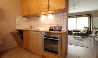 A modern kitchen with wooden cabinets and an oven. In the background, there is a living area with a cozy sofa.