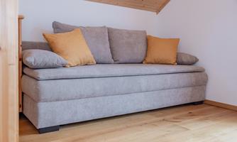 A cozy gray sofa with two pillows in warm tones. It is located in a quiet room with wooden flooring and a sloping ceiling.